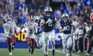 CSU BYU football player in blue jersey carrying ball, sprinting toward field with crowd in blue stands behind him.