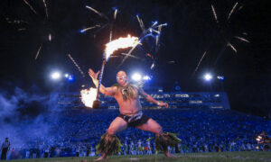 Fire dancer performing with a flaming staff on a stadium field at night, cheering crowd in background.