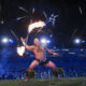 Fire dancer performing with a flaming staff on a stadium field at night, cheering crowd in background.