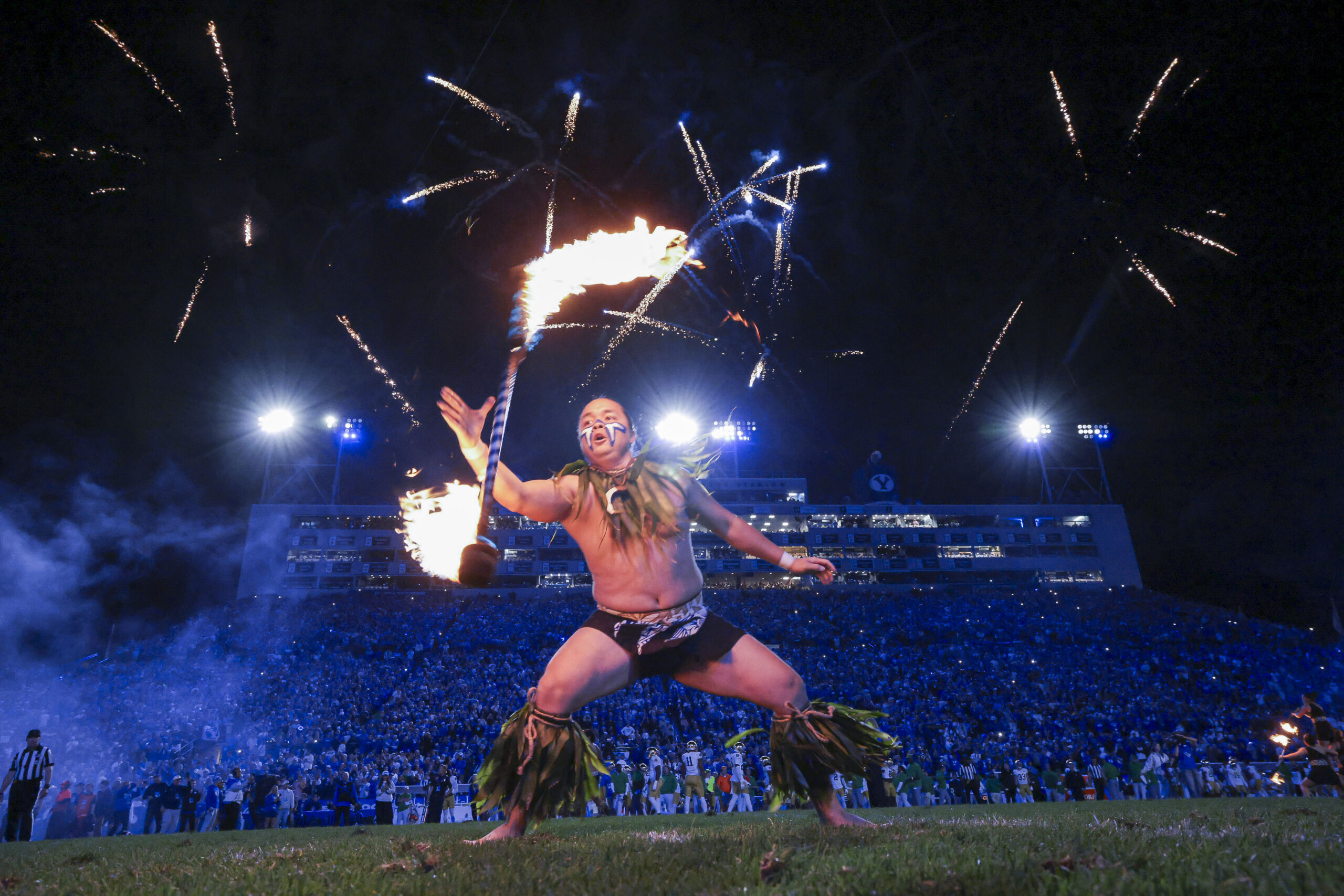 Fire dancer performing with a flaming staff on a stadium field at night, cheering crowd in background.